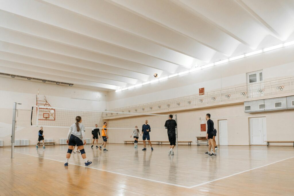 Group of athletes playing volleyball in an indoor sports hall with a wooden floor.